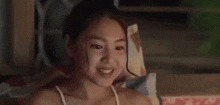 a young girl in a white tank top is smiling and sitting on a bed .