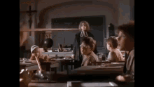 a group of children are sitting at their desks in a classroom with a teacher standing in the background .