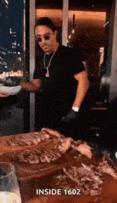 a man wearing sunglasses and a necklace is standing in front of a cutting board with steaks on it .