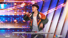 a man is singing into a microphone while standing in front of an ironing board on a stage sponsored by nbc