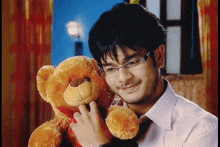 a man wearing glasses holds a teddy bear with a red ribbon around its neck