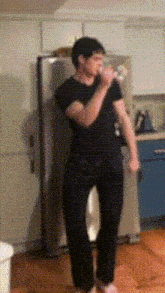 a man in a black shirt is drinking from a bottle in a kitchen