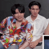 two young men are sitting next to each other in a room . one of the men is wearing a colorful shirt .