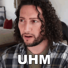 a man with curly hair and a beard is wearing a plaid shirt and saying uhm