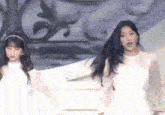 two women in white dresses are standing next to each other on a stage in front of a wall .