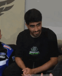 a man wearing a black shirt with a green circle on it sits on a couch