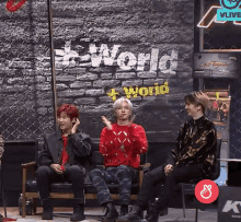 three men are sitting in front of a brick wall that says world