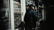 a man in a black jacket stands in front of a store with graffiti on the wall that says " again "