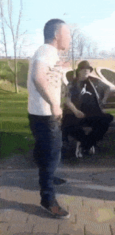 a man in a white shirt is standing next to a man in a black shirt sitting on a bench .