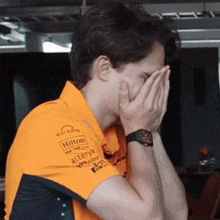 a man covering his face with his hands while wearing an orange shirt with hilton alteryx and workday written on it