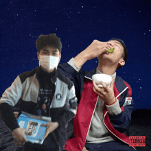 a man wearing a mask is eating a salad next to another man wearing a jacket with a ny logo on it