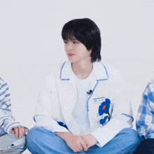 a young man wearing a white shirt with a blue flower on it sits on the floor