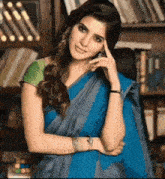 a woman in a blue saree is standing in front of a bookshelf with her hand on her forehead .
