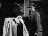 a man in a suit and hat is standing next to a woman in a fur coat in a black and white photo .