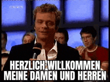 a man is holding a microphone in front of a crowd and says herzlich willkommen meine damen und herren