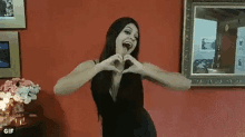 a woman in a black dress is making a heart with her hands in front of a red wall .
