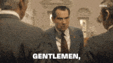 a man in a suit and tie says " gentlemen " in front of two other men