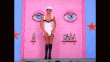 a woman in a white dress stands in front of a pink wall with large blue eyes