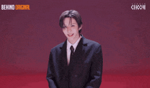 a man in a suit and tie is standing in front of a sign that says " behind original "