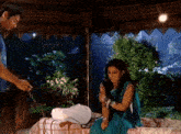 a woman in a blue sari sits on a bed while a man stands nearby
