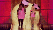 two women in pink dresses are standing on a stage holding feathered umbrellas .