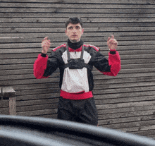 a man in a red white and black jacket stands in front of a wooden wall with his hands in the air