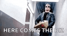 a man in a leather jacket is carrying a box in a hallway and says `` here comes the biz '' .