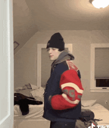 a man wearing a beanie and a varsity jacket is standing in front of a bed