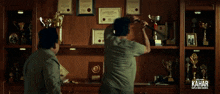two men are standing in front of a wall full of trophies and certificates and the word kahar is on the bottom right