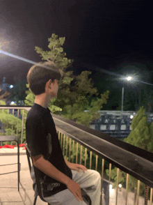 a young man sits on a balcony at night wearing a black shirt that says " you " on the back