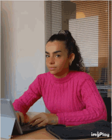 a woman in a pink sweater sits at a desk with a laptop