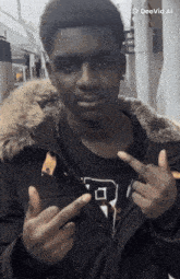 a young man wearing a black jacket with a fur hood is pointing his fingers at the camera