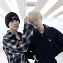 a man wearing a black stussy beanie stands next to another man wearing a plaid shirt