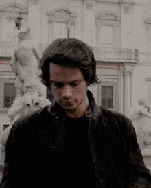 a man in a black jacket is standing in front of a building and a statue .