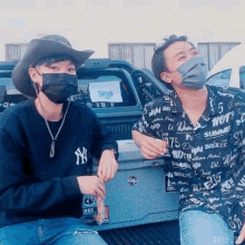 a man wearing a ny shirt sits next to another man wearing a black mask