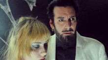 a man with a beard and earrings is standing next to a woman with blonde hair