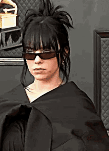 a woman wearing sunglasses and a black jacket is standing in front of a wall .