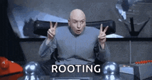 dr. evil is giving a peace sign while sitting at a desk in a room .