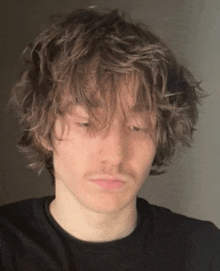 a young man with long curly hair and a mustache is wearing a black shirt .
