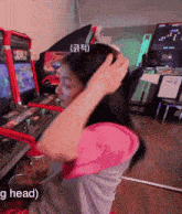 a woman adjusts her hair while playing a video game in an arcade with the words ' g head ' below her