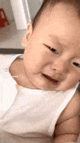 a baby in a white tank top is crying while looking at the camera .