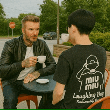 a man wearing a miu miu laughing boy t-shirt is sitting at a table with another man