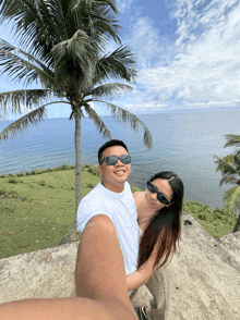 a man and woman wearing sunglasses pose for a picture in front of a palm tree overlooking the ocean
