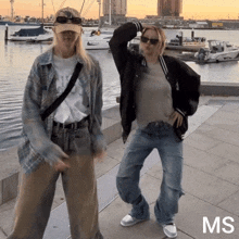 two women are dancing in front of a body of water and the word ms is on the sidewalk