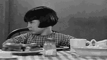 a black and white photo of a young girl sitting at a table with plates of food .