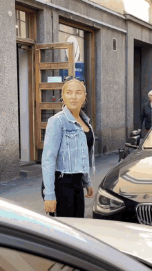 a woman in a denim jacket is standing next to a black car