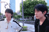 two young men are standing next to each other on a sidewalk and one of them is wearing a white jacket with black stripes ..