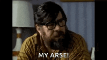 a man with a beard and glasses is sitting in front of a lamp and saying `` my arse '' .