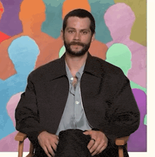 a man with a beard is sitting in a chair in front of a colorful painting