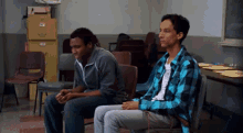 a man in a blue plaid shirt sits next to another man in a classroom